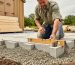 Homeowner kneeling next to a leveled gravel and concrete block shed foundation while using a level and tape measure, with a shed kit nearby in the yard.